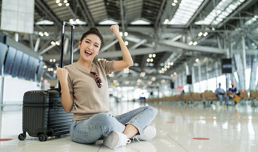 An EB5 investor sitting at an airport next to her luggage, smiling with her arms in the air.