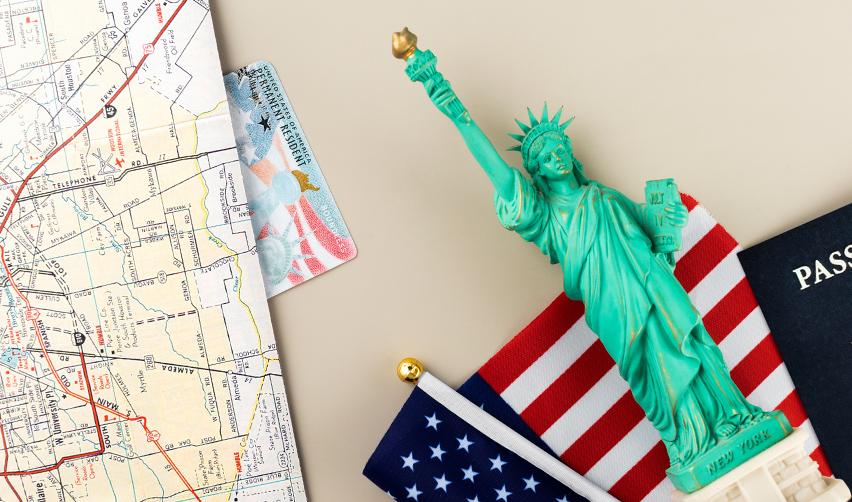 The statue of liberty, a passport, a U.S. map, a permanent resident card (green card) and the American flag on a desk.