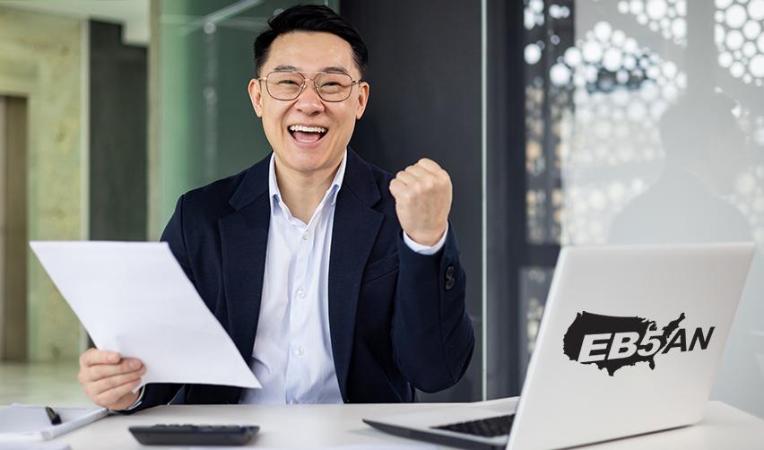 A happy alien investor holding a document in front of a computer with an EB5AN logo on it.