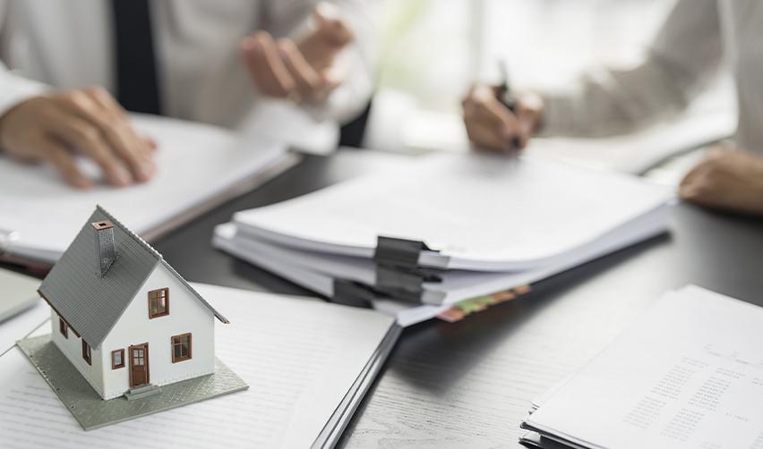 Hands of foreign investors signing some documents with a wooden house figure on a desk, symbolizing a real estate investment.