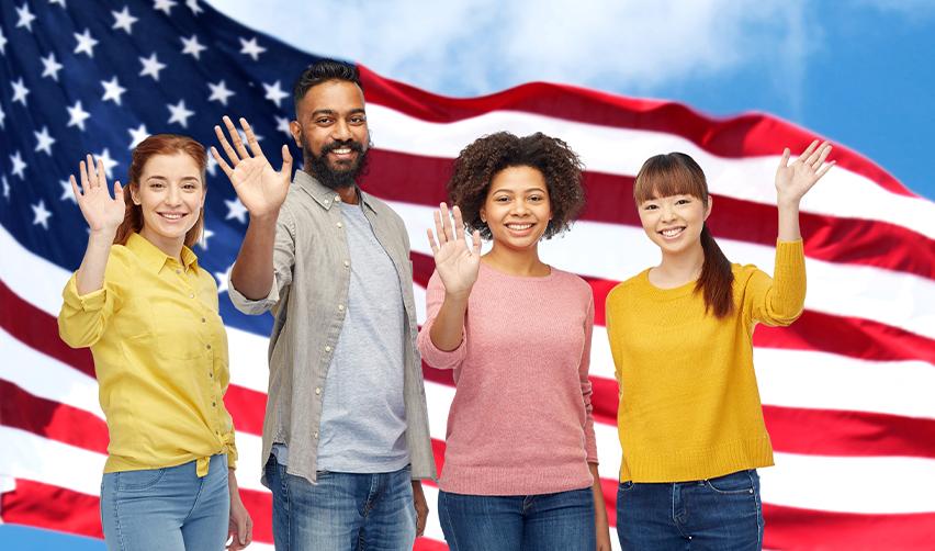 A group of foreign nationals waving at the camera with the U.S. flag behind them.