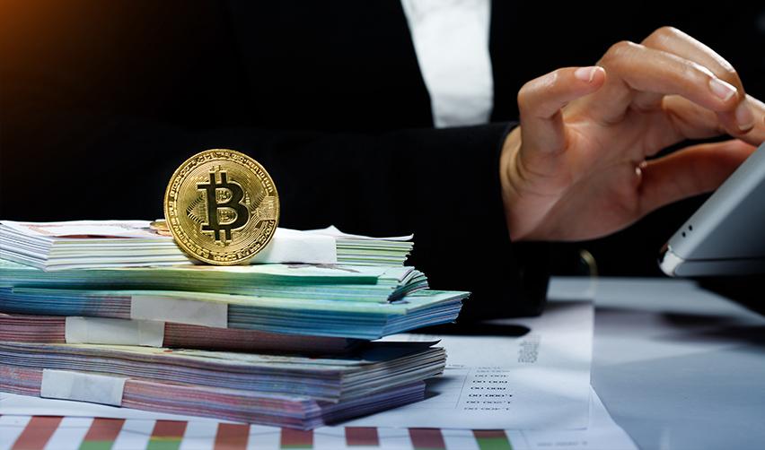 A bitcoin placed on top of stacks of money, next to an EB5 investor's hand going through their visa application process.
