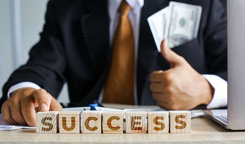 A businessman in a suit showcasing success with wooden blocks spelling "SUCCESS" and a stack of dollar bills in his pocket. This image represents financial achievement through EB5 investments.