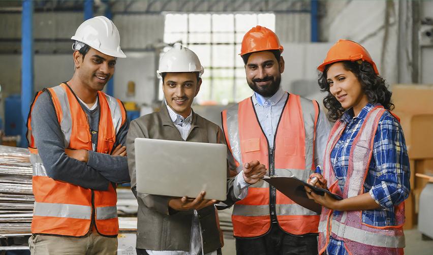 Group of Indian professionals in hard hats reviewing a laptop in a warehouse, symbolizing H1B visa workers in America.