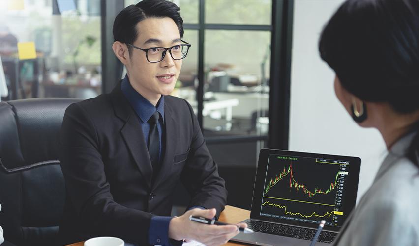 A professional financial advisor explaining EB5 investment strategies to a client with a stock market graph displayed on a laptop.