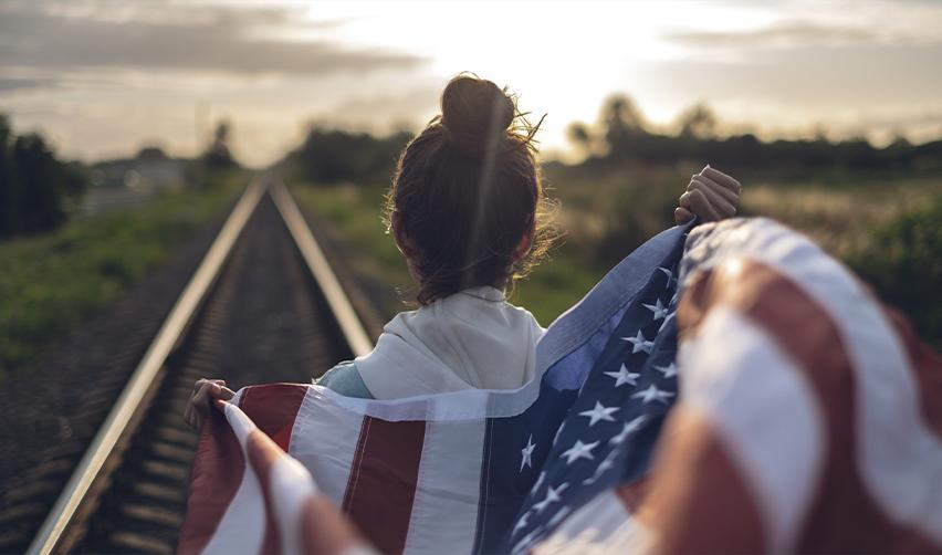 A woman holding an American flag on a railway track at sunset, symbolizing the journey toward permanent residency and the American Dream through the EB5 program.