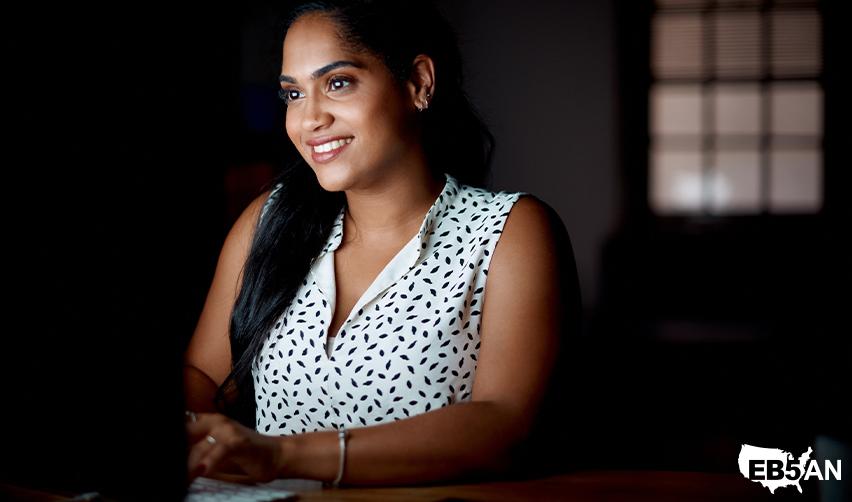The EB5AN logo next to a confident Indian businesswoman working late at her desk, highlighting the opportunities the EB5 program provides for entrepreneurs expanding their businesses in the U.S.