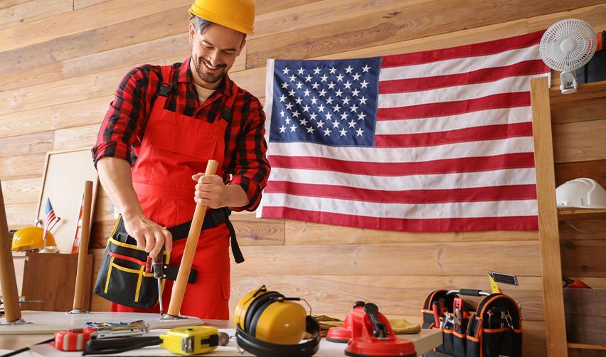 An EB-5-funded construction worker assembling furniture in a workshop, wearing a red plaid shirt and tool belt, with an American flag in the background. EB5 investments support job creation in U.S. manufacturing and skilled trades.