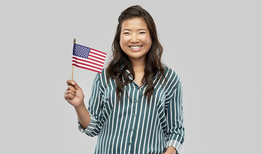 A smiling Asian woman holds a small American flag, symbolizing successful immigration to the United States through the EB5 visa program. The image conveys optimism and achievement in securing U.S. permanent residency.