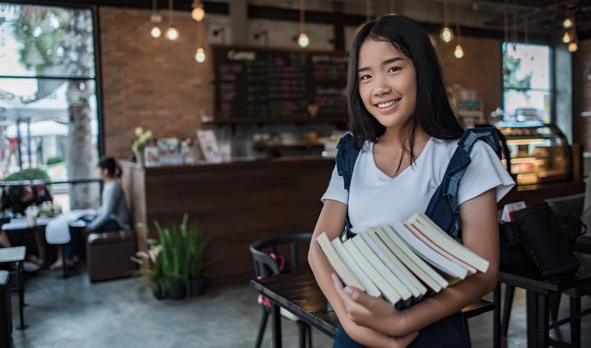 A smiling international student in a café, holding a stack of books, illustrating how F1 visa holders can finance their EB5 investment and secure a U.S. Green Card while completing their studies.