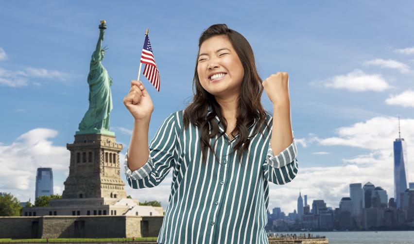 A happy Taiwanese national holding up the American flag in front of the statue of liberty, symbolizing how green card applications to the united states citizenship and immigration services under the eb5 visa can benefit citizens of taiwan.