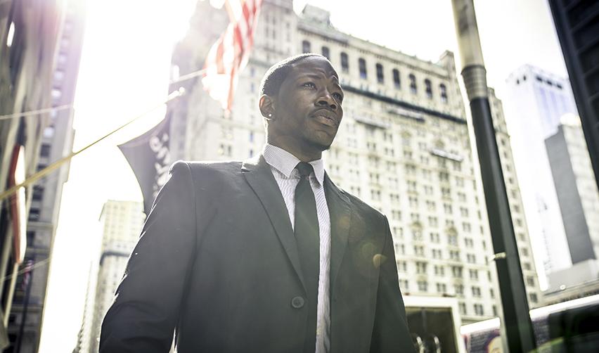 Professional man standing in a U.S. city with American flags in the background, representing international investors seeking U.S. residency through EB5 and wealth-based immigration programs.