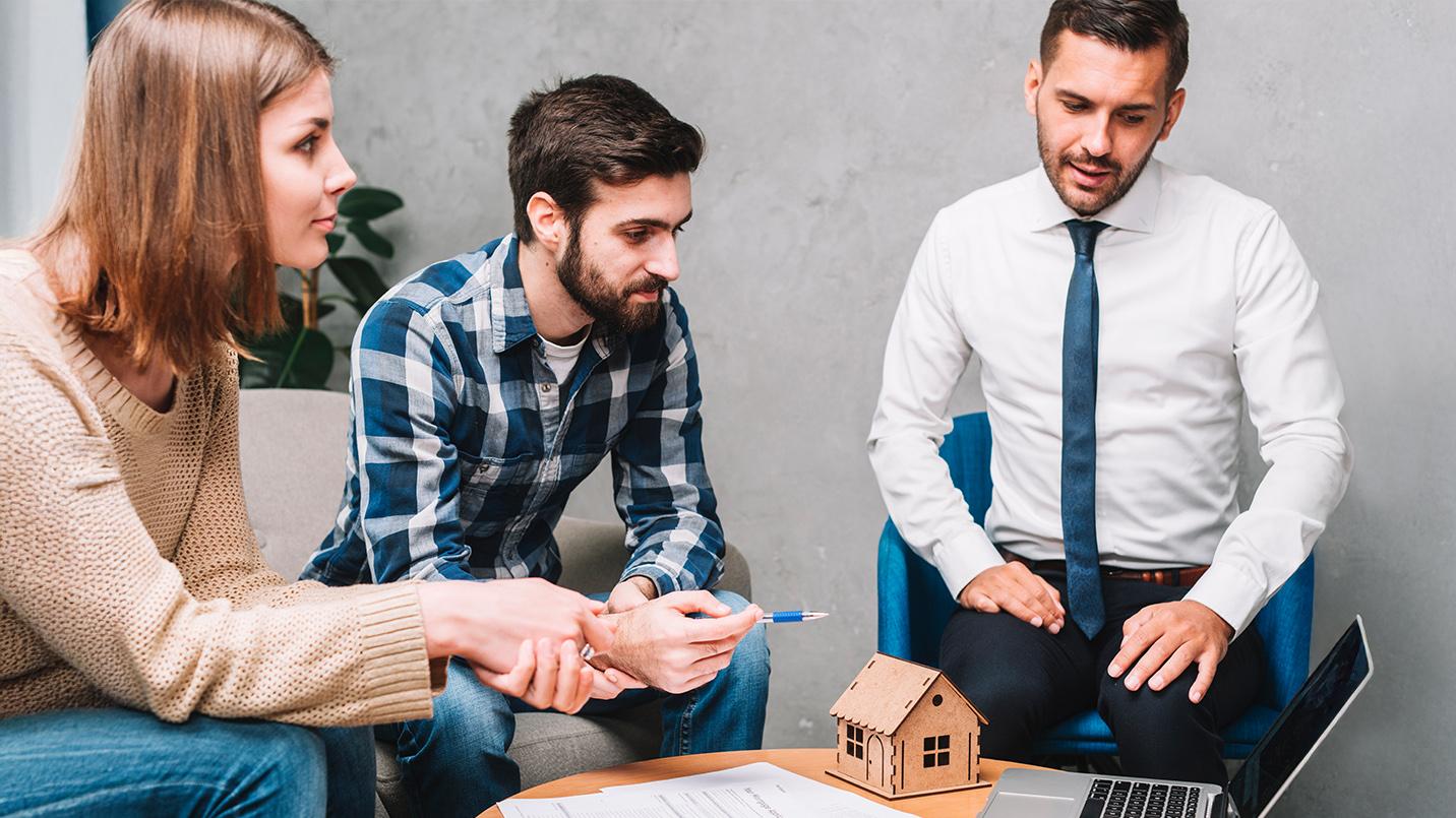 A young couple discussing real estate options with a professional advisor from PRelocate, evaluating the decision to rent or buy in Puerto Rico. The small wooden house model on the table symbolizes property investment and homeownership opportunities on the island.
