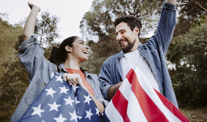 A smiling couple holding a U.S. flag outdoors, symbolizing EB5 derivative beneficiaries celebrating Green Card approval.