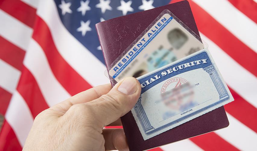 Close-up of a U.S. Resident Alien card and Social Security card held against an American flag, symbolizing U.S. residency through EB5 investment with the help of EB5AN.