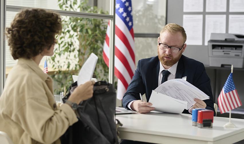 An EB5 visa applicant meeting with a consular officer at a US embassy, reviewing immigration documents with American flags in the background.