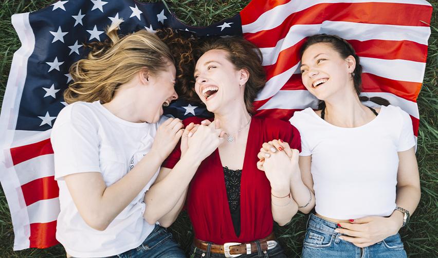 Three young women lying on an American flag outdoors, smiling and holding hands, representing community and new opportunities through the EB5 program.