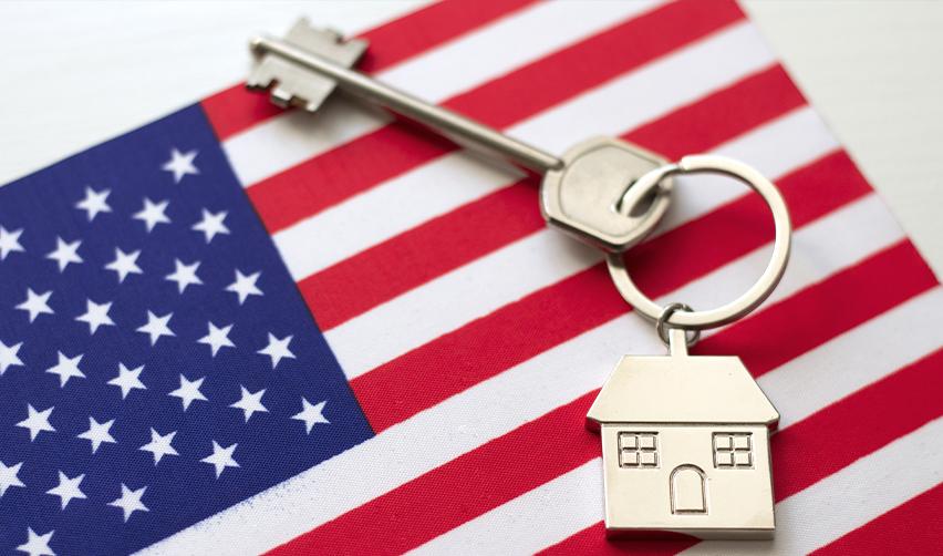 A silver house key with a home-shaped charm resting on a U.S. flag, symbolizing property ownership opportunities for EB5 investors before Green Card approval.