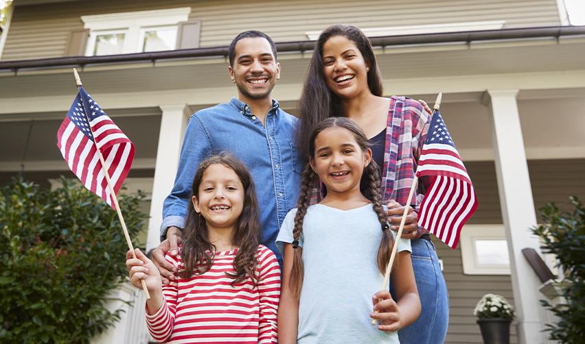 A happy immigrant family holding American flags in front of their new home, symbolizing the benefits of EB5 Green Cards and U.S. residency.