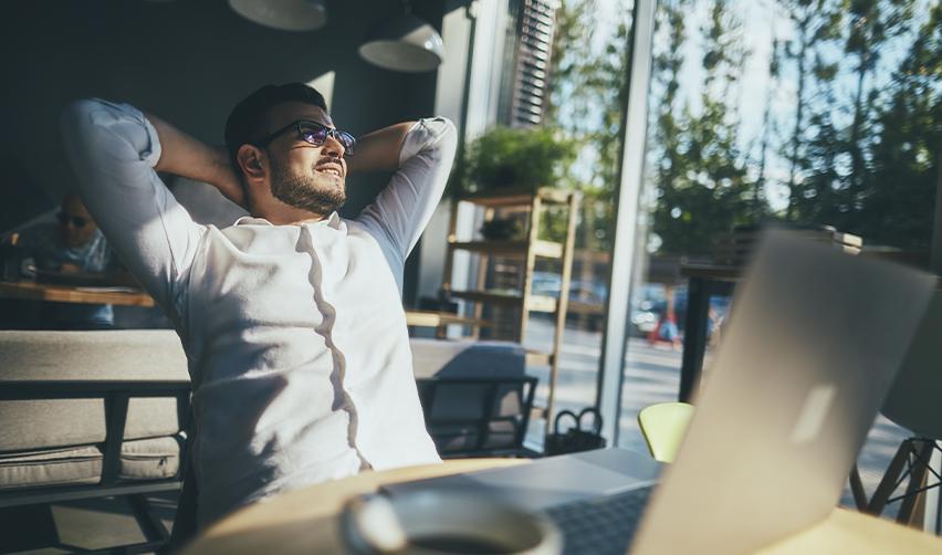 A relaxed man stretching back in front of a laptop in a sunlit café, portraying the freedom and stability EB5 investors can achieve after securing permanent residency.