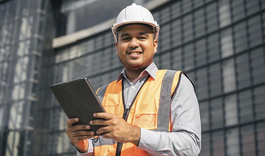 Engineer in a safety vest and hard hat using a tablet outside a building, representing H1b visa workers starting the EB5 visa process.