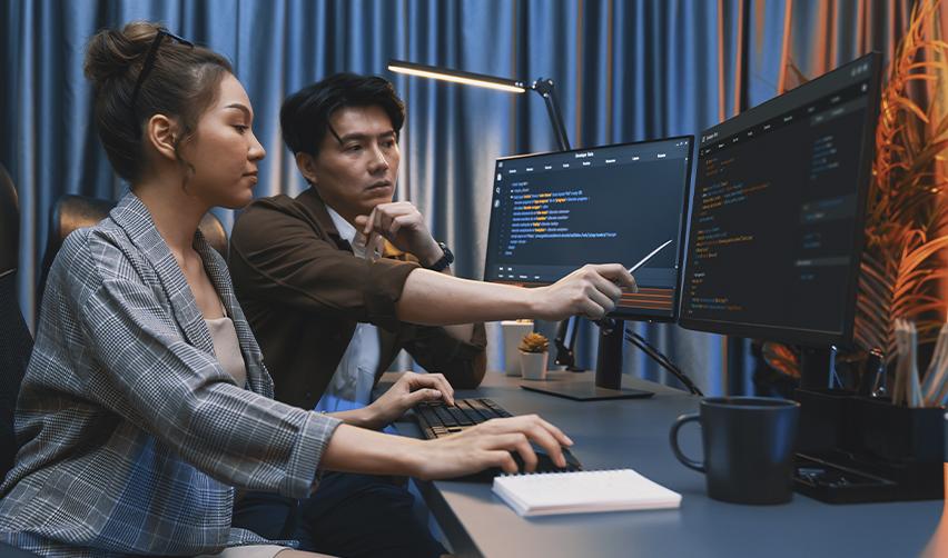 A focused pair of developers reviewing lines of code on dual monitors in a dimly lit office, representing the career uncertainty many H1B visa holders in tech face.