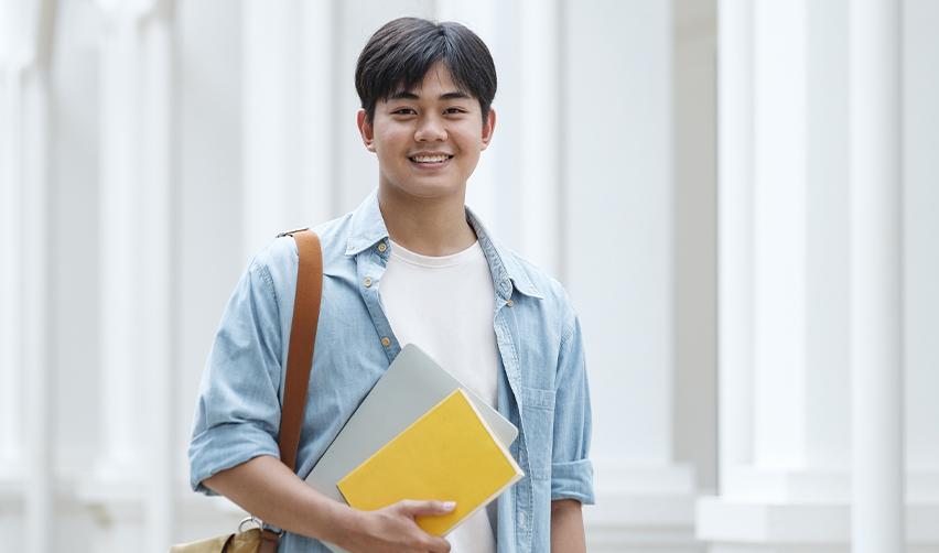 Young male student holding books and a laptop on a U.S. university campus, symbolizing EB5 families timing their investment for college education benefits.