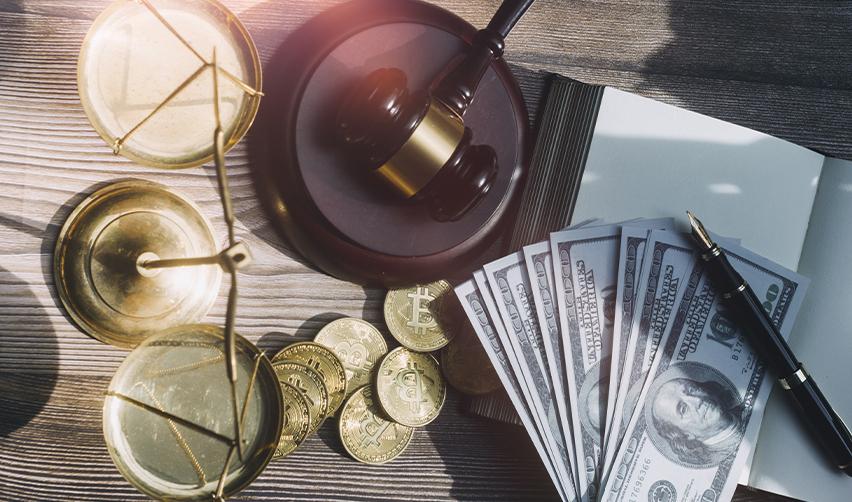 Close-up of a gavel, scales of justice, U.S. dollar bills, and gold Bitcoin coins on a wooden desk, symbolizing EB5 investment loans and lawful source of funds compliance.