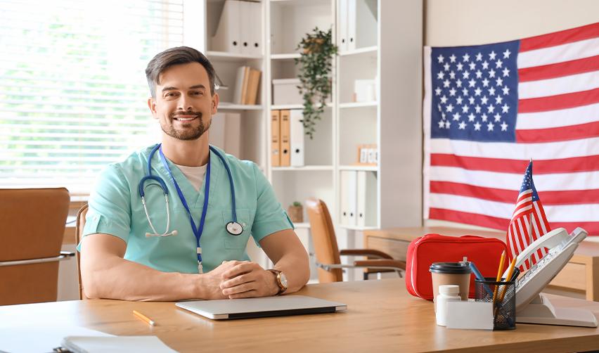 A smiling doctor seated at a desk with a U.S. flag in the background, symbolizing successful immigration and career opportunities through the EB-5 visa for doctors in USA.