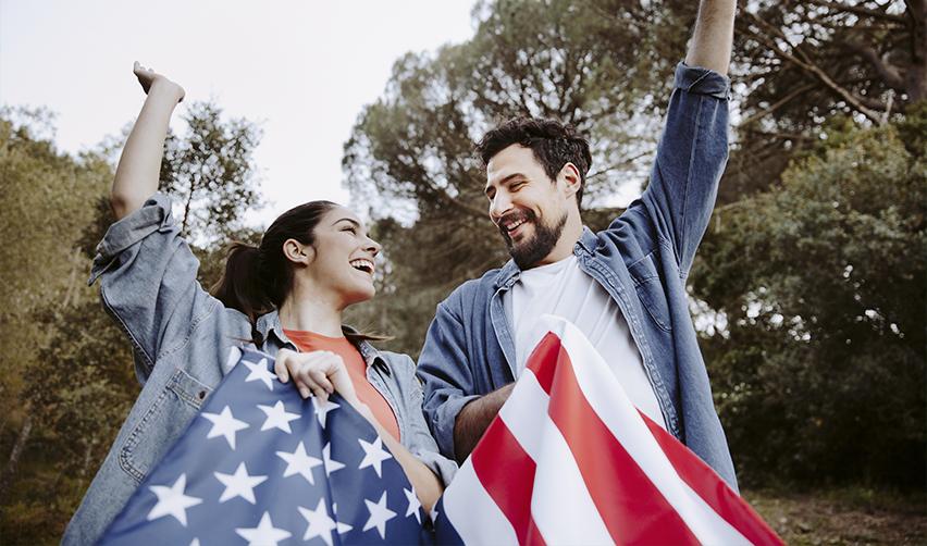 Happy couple celebrating with U.S. flags in nature, reflecting the family and lifestyle benefits of EB5 immigration.