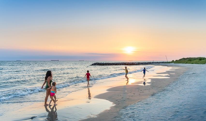 A family walks and plays along the shoreline of Cape Charles Beach at sunset, with golden light reflecting on the sand and waves of Chesapeake Bay.