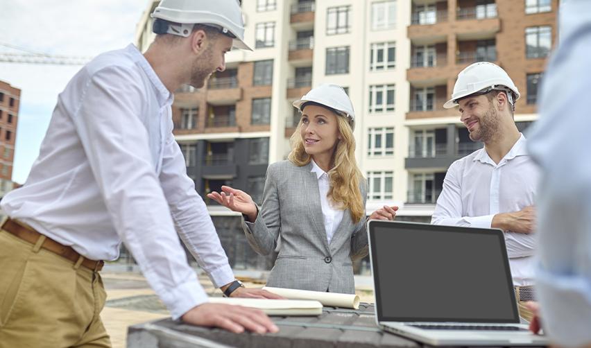 Developers and architects in hard hats discussing plans at a construction site with laptops and blueprints, reflecting teamwork in EB5 real estate development.
