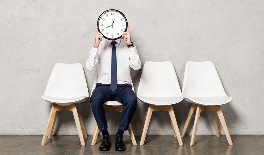 A man in a business suit sitting in a waiting area holding a clock in front of his face, symbolizing EB5 investors anticipating visa processing and approval timelines.