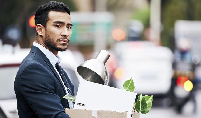 A laid-off H1B worker in a suit carrying a box with office items after losing his job, symbolizing visa uncertainty for foreign workers facing layoffs in the U.S.