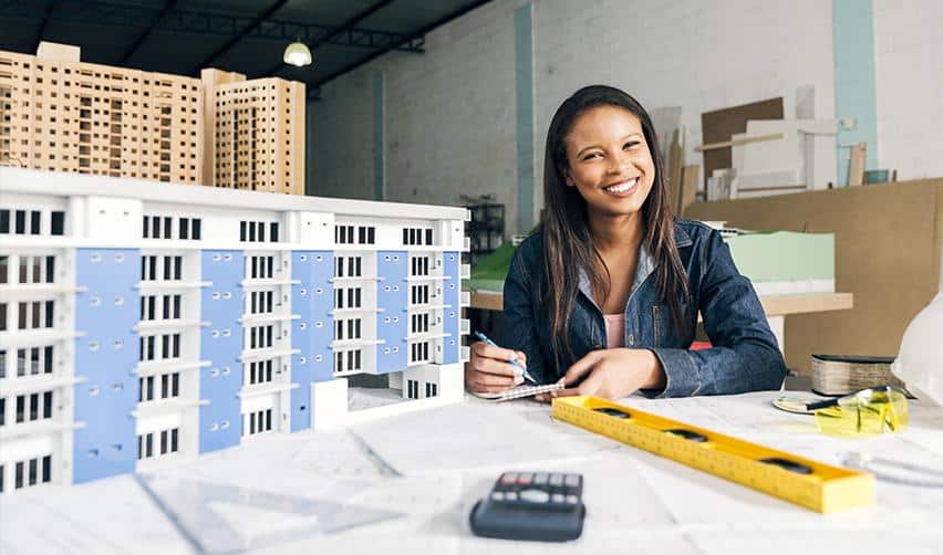 Architect smiling beside a scale model of an apartment complex, symbolizing planning, design, and construction roles supported by EB5 real estate projects.