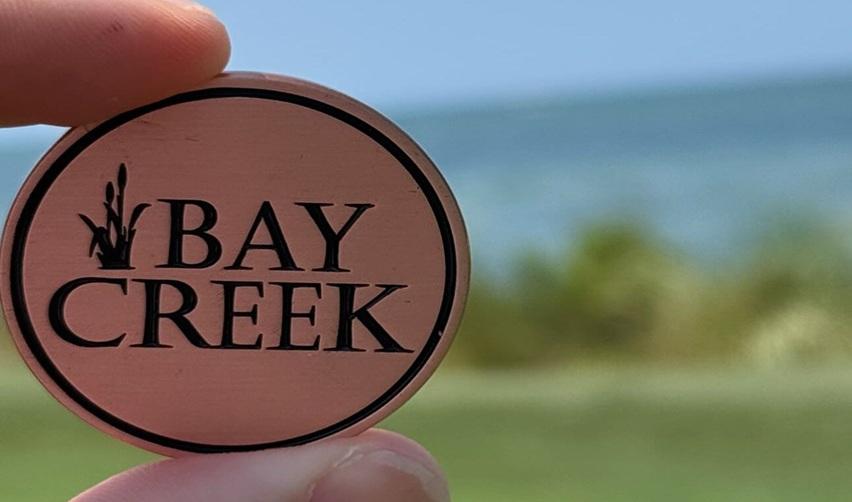 Hand holding a Bay Creek token against the backdrop of the Chesapeake Bay shoreline, representing the community’s brand and the value of EB5-backed development.