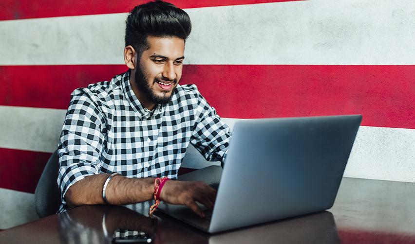 Young Indian professional working on a laptop in front of an American flag wall, representing EB5 as a pathway to U.S. residency beyond H1B limitations.