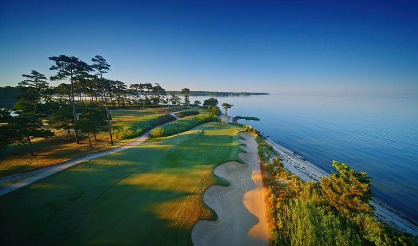 Aerial view of Bay Creek’s waterfront golf course along the Chesapeake Bay, showing greens bordered by sand bunkers, native vegetation, and calm coastal waters, symbolizing the community’s sustainable design and EB5 investment appeal.