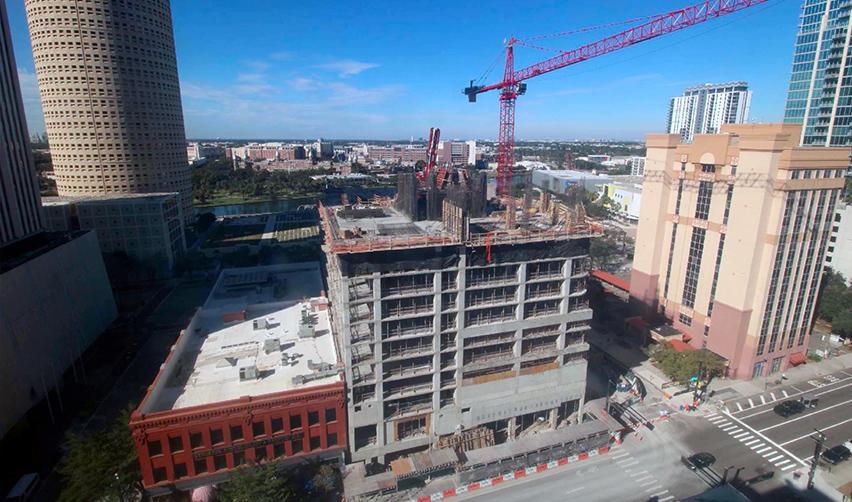 Construction progress at ONE Tampa showing early-stage vertical work, concrete framework, and tower cranes rising above downtown Tampa under clear blue skies.