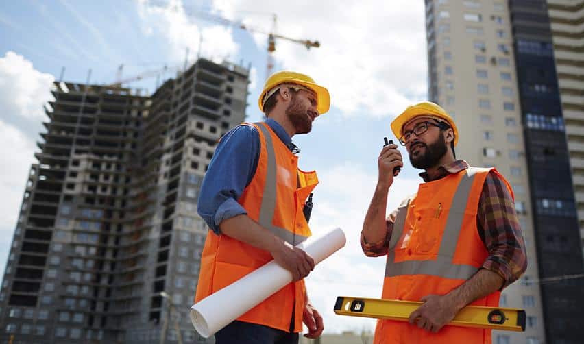 Two construction engineers wearing orange safety vests discussing plans at a building site, reflecting job creation and project development under the EB5 program.
