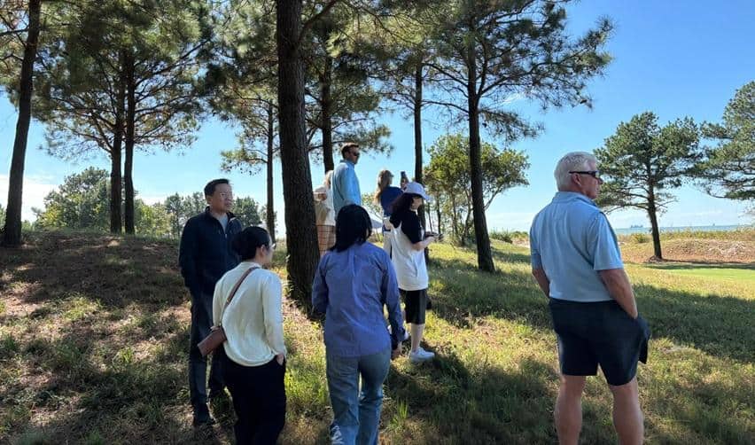 A small group of EB5AN site visit attendees exploring Bay Creek’s nature preserve area, standing among pine trees overlooking the Chesapeake Bay on a sunny afternoon.