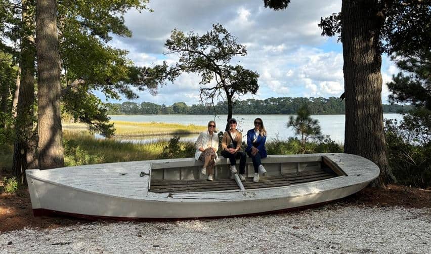 Three EB5AN site visit guests seated on a beached wooden rowboat by the Bay Creek shoreline, with calm waters and pines in the background reflecting the area’s natural beauty.