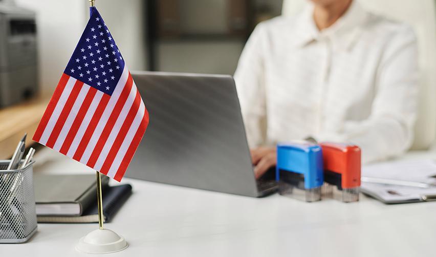 A desk with a small US flag, office supplies, and a person working on a laptop, symbolizing EB5 visa applications and immigration paperwork.
