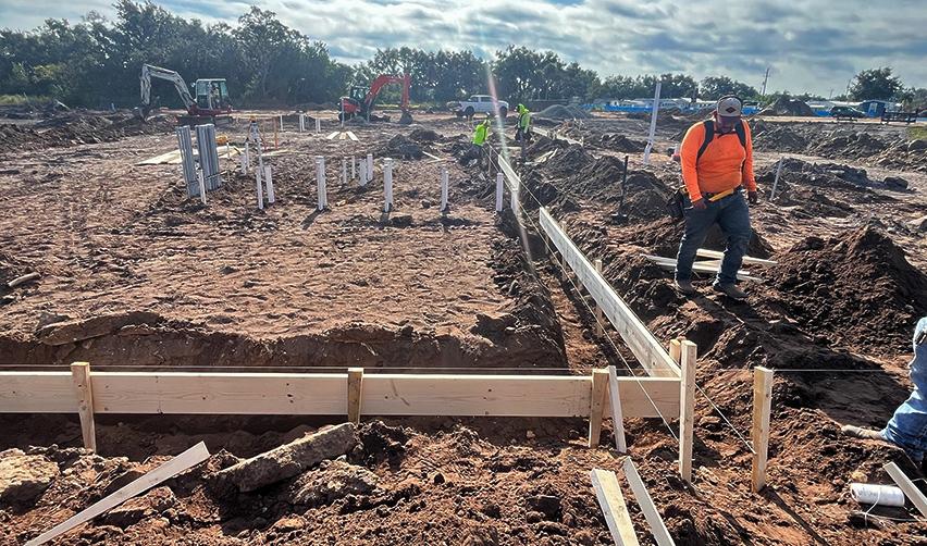 Construction workers preparing foundation trenches at the Terra Ceia Multifamily EB5 site, with plumbing lines, framing boards, and excavators visible across the active work area.