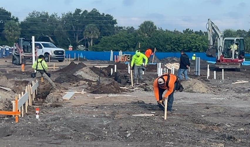 Workers marking layout lines and installing utility piping across the Terra Ceia Multifamily EB5 construction site, with excavators and trucks operating in the background.