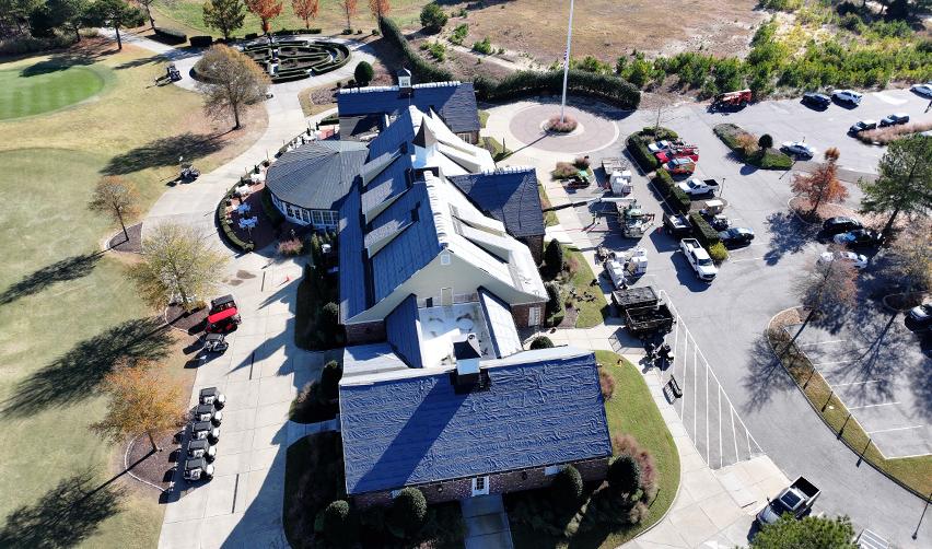 Overhead photo of the Bay Creek Clubhouse roof showing active shingle installation and construction equipment positioned around the building.