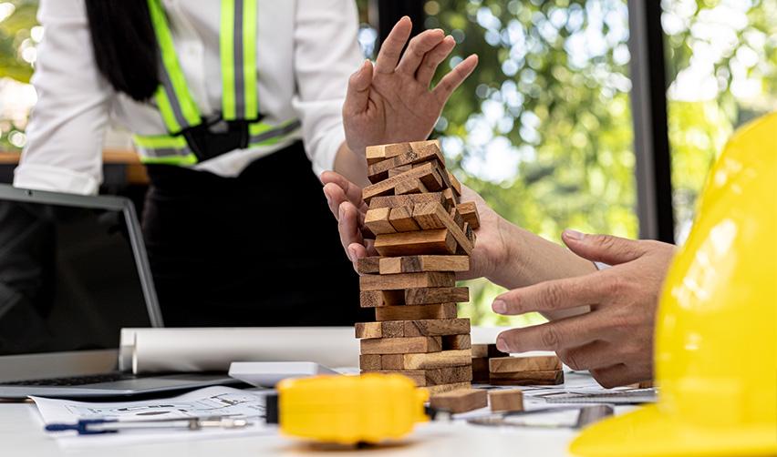 Hands carefully stabilizing a stack of wooden blocks on a worktable with construction plans and safety gear, symbolizing how complexity and instability can increase risk in EB5 investment project structures.