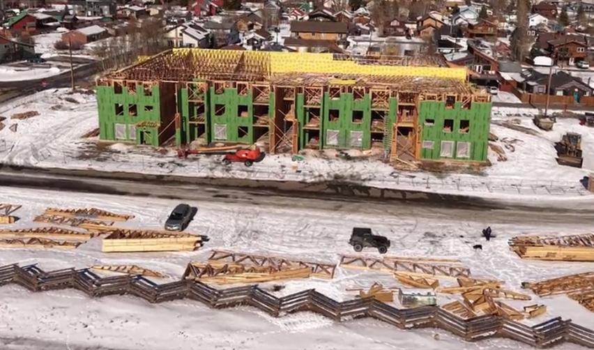 Aerial view of a multi-story building under construction at the Grand Park community, showing green exterior sheathing, roof framing in progress, and active winter construction supporting EB5 job creation in this rural Colorado project.