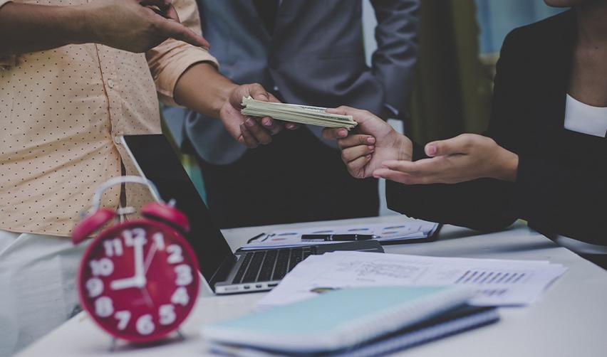 Hands exchanging cash over a desk with financial documents and a clock, symbolizing EB5 fund deployment timing, capital release, and sustainment considerations in EB5 investment structures.