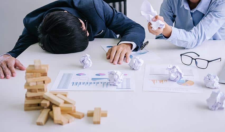 A toppled stack of wooden blocks on a desk beside financial charts and stressed professionals, illustrating project failure and hidden risks that EB5 investors may face in overly complex or poorly structured investments.